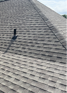 A close-up view of a house roof covered with gray asphalt roofing shingles, showing a vent pipe protruding near the roof peak under a partly cloudy sky.