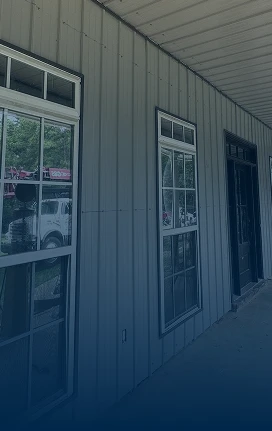 A building with gray metal siding and roofing, large rectangular windows, and a black door; a white truck is partially visible through the window reflection.